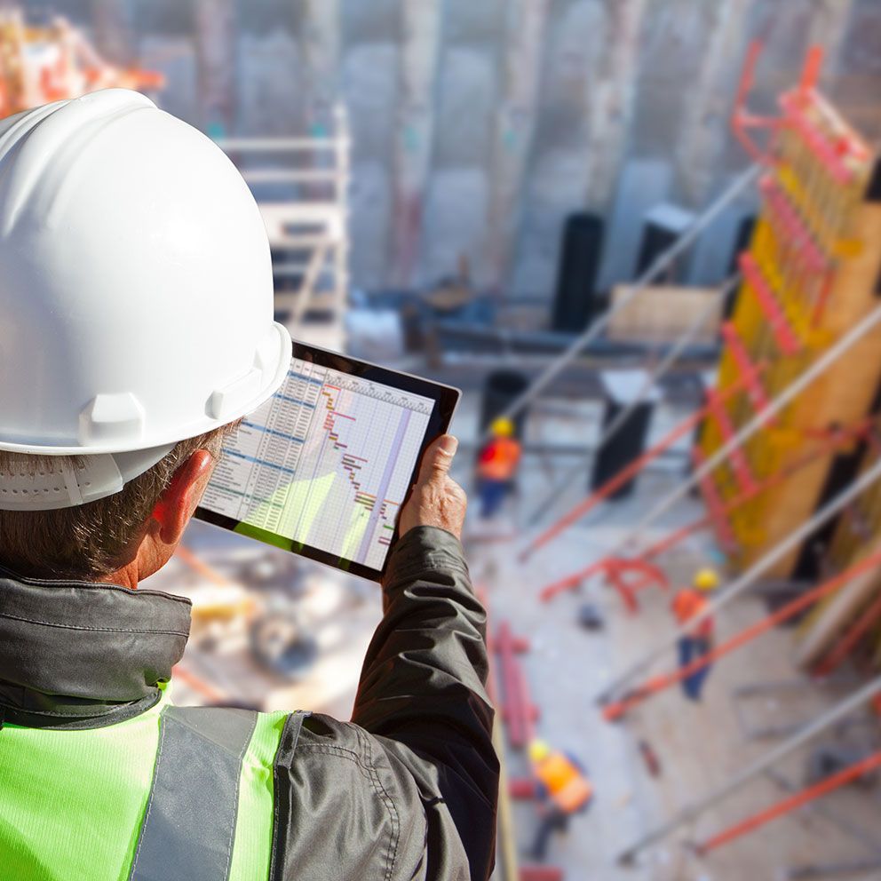 Bauarbeiter mir Helm steht mit dem Rücken und einem iPad in der Hand vor einer Baustelle